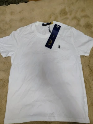 Vit t-shirt från Polo Ralph Lauren - Klassisk vit t-shirt från Polo Ralph Lauren med rund halsringning och ikoniska broderade loggan på bröstet. Tillverkad i mjuk bomull för en skön känsla. Perfekt basplagg med stilren design och diskret branding.