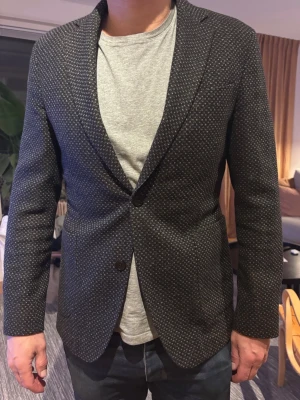 Svart kavaj med diskret mönster - Stilren svart kavaj med små vita prickar och klassisk passform. Kavajen har smala slag, enkelknäppning med en knapp och två fickor framtill. Perfekt för en dressad look och passar till både jeans och skjorta. Slim 50 men passar en 48 bättre. 