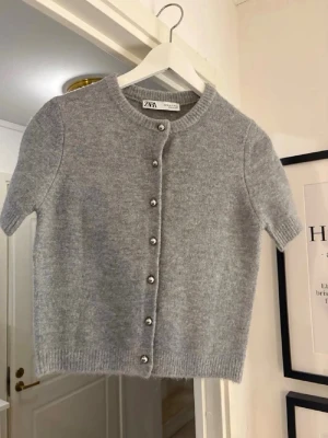 Grå kortärmad kofta från Zara - Söt grå kofta från Zara med korta ärmar och rund hals. Koftan har pärlknappar framtill och är stickad i ett mjukt material. Perfekt att slänga över en topp för en chill och stilren look.
