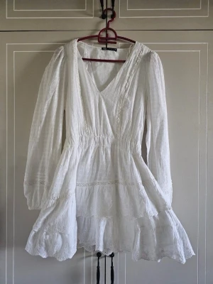 Vit omlottklänning med volang - Superfin vit kortklänning med omlott och v-ringning. Klänningen har långa ärmar, volangdetaljer och spetsinslag som ger en romantisk känsla. Perfekt för varma dagar och somriga tillfällen.