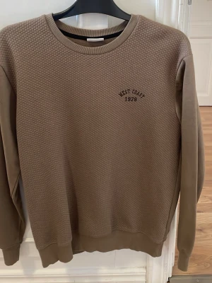 Brun sweatshirt med text West Coast - Säljer en brun sweatshirt med strukturmönstrad framsida och broderad text 'West Coast 1978' på bröstet. Tröjan har rund halsringning, ribbade muddar och är tillverkad i mjukt material. Perfekt för en avslappnad stil.