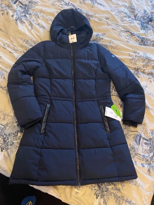 Mörkblå lång pufferjacka från Esprit - Snygg mörkblå pufferjacka från DKNY med huva och dragkedja framtill. Jackan är lång, har två fickor med svarta detaljer och quiltad design. Perfekt för kalla vinterdagar och riktigt varm tack vare det tjocka materialet.Helt ny med etikett 