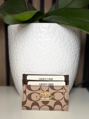 Coach brun korthållare i läder - Snygg korthållare från Coach i brunt läder med klassiskt monogrammönster och guldfärgad logga framtill. Perfekt för att organisera dina kort på ett stilrent sätt. Smidig och tunn design som enkelt får plats i fickan eller väskan.