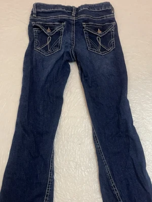 Mörkblå bootcut jeans med fickdetaljer - Säljer ett par mörkblå bootcut jeans med snygga kontrastsömmar och dekorativa fickor bak med knapp och broderade detaljer. Klassisk femficksmodell och låg midja. Perfekta för dig som gillar en avslappnad men trendig stil.