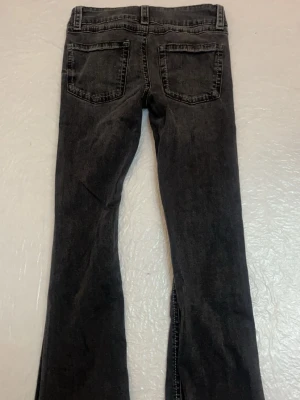 Svarta bootcut jeans med dubbla knappar - Säljer ett par svarta bootcut jeans med dubbla knappar i midjan och fem fickor. Jeansen har en lätt tvättad look och utsvängda ben. Snygga detaljer vid fickorna framtill och klassisk jeanssöm. Perfekta för en trendig och avslappnad stil.