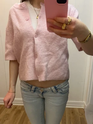 Ljusrosa ribbad kofta med knappar - Kort, ljusrosa kofta med ribbad struktur med knappar längs mitten. Koftan har halvlånga ärmar och knäpps framtill med matchande knappar. Perfekt att slänga över ett linne eller t-shirt för en chill look.