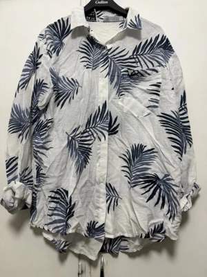 Vit skjorta med blå palmbladsmönster  - Snygg vit skjorta med blå palmbladsmönster. Skjortan har klassisk krage, knäppning framtill och långa ärmar som kan vikas upp. Tillverkad i ett lätt och luftigt material, perfekt för en avslappnad stil.