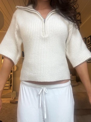 Vit ribbad stickad tröja med dragkedja - Vit stickad tröja med ribbad struktur och halv dragkedja framtill. Tröjan har korta, vida ärmar och en liten krage som kan vikas ner. Materialet ser mjukt och stretchigt ut, perfekt för en avslappnad men ändå snygg look.