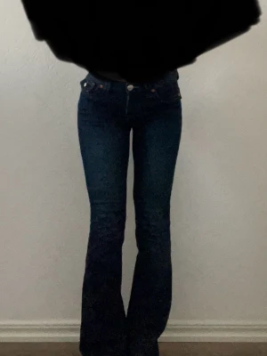True Religion Becca Bootcut Jeans - Säljer ett par mörkblå True Religion Becca jeans med bootcut och snygga kontrastsömmar. Modellen har mellanmidja och klassiska fickor med ikoniska detaljer och silverknappar. Perfekt passform och riktigt schysst tvätt.
