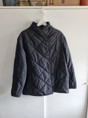 Arket svart quiltad jacka S oversize - Stilren svart pufferjacka med quiltat mönster och sned knäppning framtill. Jackan har en hög krage och tryckknappar, vilket ger en modern look. Perfekt för kalla dagar och enkel att matcha med olika outfits. Sparsamt använd, perfekt skick.
