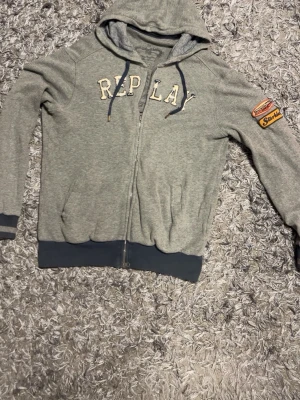 Replay Hoodie- strl M - Grå hoodie med dragkedja från Replay. Stor logga i vitt och beige på bröstet och patch på ena ärmen. Mörkgrå muddar vid ärmslut och nederkant samt två ränder på ena ärmen. Huva med dragsko och fickor fram. Skön och avslappnad stil, perfekt till jeans.