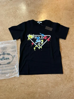 Svart Prada t-shirt med färgstänk - Snygg svart t-shirt från Prada med ikoniskt Milano-tryck och färgstänk i gult, blått och rött framtill. Klassisk passform och rund halsringning. Storlek small, passar någon i storlek xs. Helt ny i förpackning 