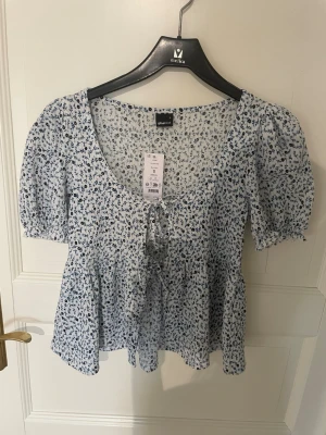 Blommig blus med knyt från Gina Tricot - Supersöt blus från Gina Tricot i vitt med blått blommönster. Modellen har korta puffärmar, v-ringning och knyt framtill.