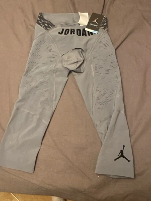 Gråa Jordan träningstights - Snygga gråa träningstights från Jordan med svart logga på benet och texten JORDAN framtill. Tightsen har ventilerande meshdetaljer och elastisk midja med mönster. Perfekta för sport och träning.