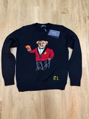 Svart stickad tröja Polo Bear - Svart stickad tröja från Polo Ralph Lauren med ikoniska Polo Bear-motivet framtill. Björnen har röd kavaj och håller spelkort. RL-broderi i gult nedtill. Rund halsringning och ribbade muddar. Perfekt för dig som gillar statement-plagg.