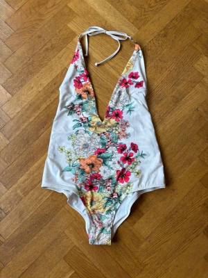Vit baddräkt med blommigt mönster - Snygg vit baddräkt med djup V-ringning och knyt i nacken. Framsidan har ett färgglatt blommigt mönster i rosa, orange, gult och grönt. Tillverkad i stretchigt material, perfekt för stranden eller poolen.