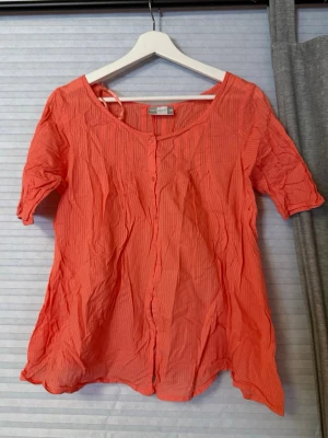 Korallfärgad blus  - Säljer en korallfärgad blus med korta ärmar och knappar framtill. Blusen har en lätt rynkad struktur och är tillverkad i ett tunt, luftigt material. Perfekt för dig som gillar färg och enkel stil.