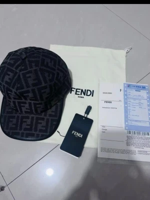 Svart keps med Fendi-logga - Säljer en svart keps från Fendi med det klassiska FF-monogrammet över hela kepsen. Kepsen har böjd skärm och justerbar rem bak. Tillverkad i ett tygmaterial och levereras med originalpåse och tagg. Perfekt för dig som vill ha en exklusiv och trendig accessoar.