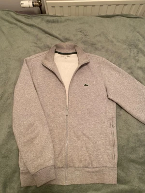 Grå ziptröja från Lacoste - Snygg grå ziptröja från Lacoste med klassisk ståkrage och dragkedja hela vägen. Tröjan har ribbade muddar och två fickor på sidorna. Ikoniska Lacoste-loggan i grönt broderad på bröstet. Perfekt för en chill och stilren look.