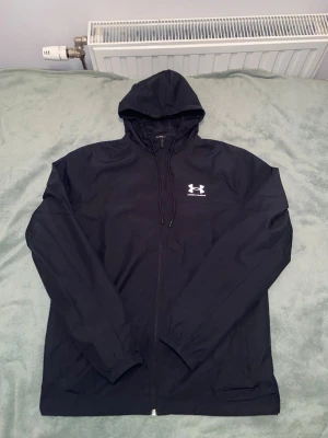 Under Armour Rival vindjacka  (ny) - Svart vindjacka från Under Armour med huva och dragkedja framtill. Jackan har vitt logotryck på bröstet och är tillverkad i lätt, vindtätt material med meshfoder. Perfekt för blåsiga dagar och sportiga outfits. Använd någon gång bara den är i stortsätt ny!
