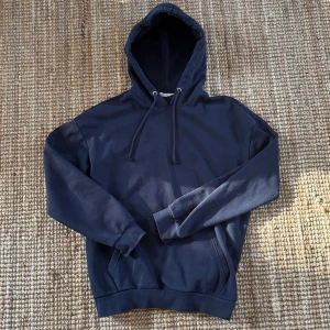 Mörkblå hoodie med ficka från pier one - En stilren mörkblå hoodie med huva och dragsnören. Hoodien har en klassisk känguruficka framtill och ribbade muddar vid ärmslut och nederkant. Perfekt för en avslappnad look och enkel att matcha med andra plagg.