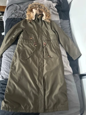 Olivgrön lång parkas med fuskpäls - Säljer en olivgrön lång parkas med mysig beige teddyfoder och huva med fuskpäls. Jackan har dragsko i midjan, två stora fickor och stängs med dragkedja och knappar. Perfekt för kalla dagar och riktigt snygg streetstil. Priset går att diskutera 