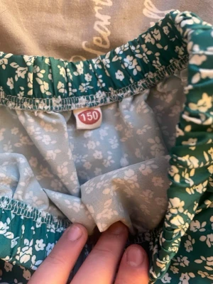 Grön blommig kjol med resårmidja - Söt grön kjol med vitt blommönster och resår i midjan. Kjolen har en luftig och lätt form, perfekt för varmare dagar. Den har en volangdetalj och är i en längre modell. Storlek 150.