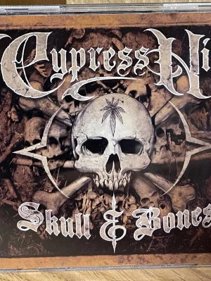 Cypress Hill Skull & Bones CD - Dubbel-CD med albumet Skull & Bones av Cypress Hill. Omslaget har en mörk design med dödskallar och ben, samt bandets logotyp i gotisk stil. Baksidan visar låtlistan och bandmedlemmar. Perfekt för dig som gillar hiphop och rock.