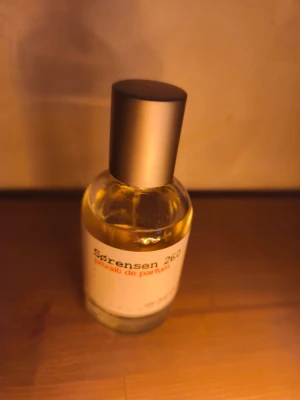 Sørensen 26.0 Extrait de Parfum - En stilren parfymflaska från Sørensen med doften 260 Extrait de Parfum. Flaskan är genomskinlig med guldfärgad vätska och har en matt svart cylinderformad kork. Etiketten är vit med svart och orange text, vilket ger en modern och minimalistisk känsla.