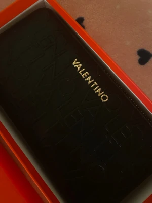 Svart plånbok från Valentino - Snygg svart plånbok från Valentino med präglad logotyp över hela framsidan och guldfärgad VALENTINO-text. Plånboken har en stilren, rektangulär form och är tillverkad i ett slätt material som ser ut som konstläder. Perfekt för dig som gillar exklusiva accessoarer.