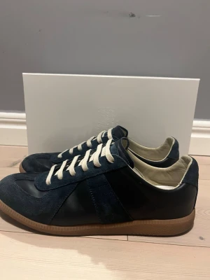 Marinblå sneakers från Maison Margiela - Snygga marinblå sneakers från Maison Margiela med ovandel i mocka och läder, vita snören och klassisk gummisula. Skorna har en låg profil och stilren design som passar perfekt till streetwear eller casual outfits.