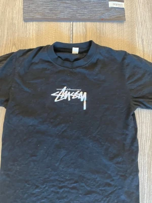 Svart Stüssy t-shirt med logga - Säljer en svart t-shirt från Stüssy med deras ikoniska vita logga tryckt på bröstet. Klassisk passform och rund halsringning. Perfekt för dig som gillar streetwear och vill ha en enkel men snygg look.