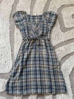 Rutig kortklänning med snörning - Kortklänning i blått, beige och vitt rutigt mönster. Klänningen har korta ärmar, snörning och knappar framtill samt ett knytband i midjan. 