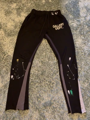 Gallery dept byxor - Säljer ett par svarta mjukisbyxor från Gallery Dept. med vitt tryck på vänster lår och färgstänk i vitt, grönt och gult på benen. Byxorna har gråa paneler längs sidorna, resår i midjan och dragsko. Perfekta för en avslappnad och kreativ stil. S men sitter som XS