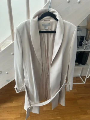 Ljusbeige kappa med skärp - Stilren ljusbeige kappa från Ivyrevel med bred krage och avtagbart midjeskärp. Kappan har en rak passform, långa ärmar och två fickor framtill. Perfekt för dig som gillar minimalistisk och trendig stil.