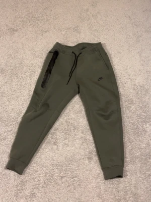 Olivgröna mjukisbyxor - Snygga olivgröna mjukisbyxor med svarta detaljer och dragsko i midjan. Byxorna har smal passform, muddar vid bensluten och en dragkedjeficka på sidan. Perfekta för chill eller träning. Passar både M och S men är lite korta om man har M