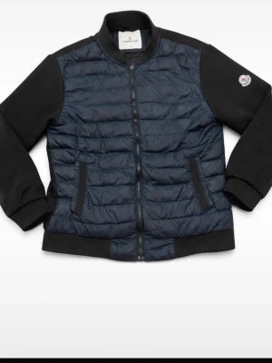 Moncler blå och svart cardigan - Snygg Cardigan från Moncler i mörkblått med svarta ärmar och muddar. Jackan har quiltad framsida, två fickor med dragkedja och klassisk Moncler-logga på ena ärmen. Materialet är en mix av nylon och bomull för en sportig look.