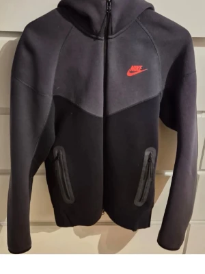Svart Nike hoodie med röd logga - Snygg svart hoodie från Nike med dragkedja framtill och två fickor med dragkedja. Den har en röd Nike-logga på bröstet och en stor huva. Tillverkad i mjukt och bekvämt material, perfekt för en avslappnad stil.
