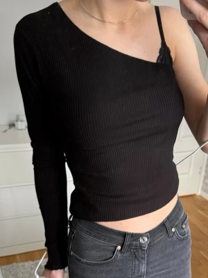 Svart oneshoulder topp från NA-KD - Trendig svart oneshoulder topp från NA-KD i ribbad bomullsmix. Croppad modell med lång ärm på ena sidan och axelbandslös på andra, perfekt för en edgy look. Snygg passform som framhäver figuren.