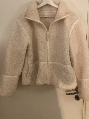 Krämvit teddyjacka med dragkedja - Mysig teddyjacka i krämvit färg med hög krage och hel dragkedja framtill. Jackan har en boxig passform och markerade sömmar som ger en cool look. Perfekt för kalla dagar när du vill vara både varm och trendig.