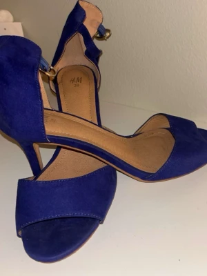 Koboltblå pumps från H&M - Snygga koboltblå pumps från H&M med öppen tå och smal klack. Skorna har en elegant ankelrem med guldfärgat spänne och är tillverkade i mockaimitation. Perfekta för att lyfta vilken outfit som helst.