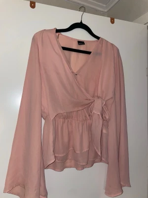 Rosa omlottblus med knytband - Söt ljusrosa omlottblus från Bik Bok med knytband i midjan och v-ringning. Blusen har långa, vida ärmar och är tillverkad i ett tunt, flowy material som ger en romantisk känsla. Perfekt att styla med jeans eller kjol för en trendig look.