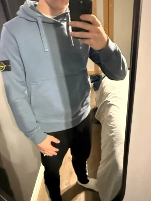 Ljusblå hoodie från Stone Island - Snygg ljusblå hoodie från Stone Island med klassisk huva och känguruficka framtill. Ikonisk logopatch på vänster ärm. Perfekt för en avslappnad streetwear-look. Mjuk bomullskänsla och ribbade muddar vid ärmslut och nederkant.