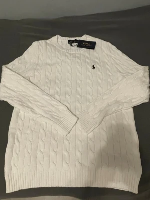 Vit kabelstickad tröja Polo Ralph Lauren - Vit kabelstickad tröja från Polo Ralph Lauren med rund halsringning och klassisk broderad logga på bröstet. Tröjan har långa ärmar och ribbade muddar. Perfekt för en stilren och tidlös look. Butikpris:1099+ Mitt pris:529