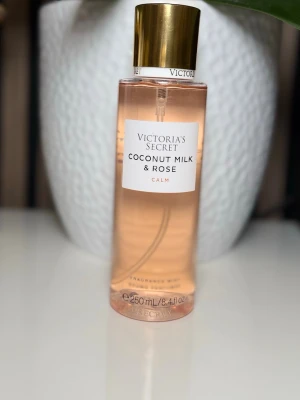 Victoria's Secret Coconut Milk & Rose - Fräsch parfym från Victoria's Secret med doft av kokosmjölk och ros. Flaskan är cylinderformad i transparent plast med ljusrosa vätska och guldfärgat lock. Rymmer 250 ml och har en elegant, modern design.