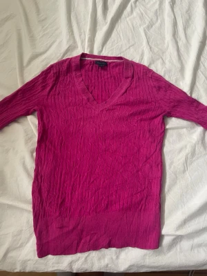 Rosa stickad tröja Tommy Hilfiger - Snygg rosa stickad tröja från Tommy Hilfiger med kabelstickat mönster och djup V-ringning. Tröjan har långa ärmar och bred ribbad mudd vid nederkant och ärmslut. Perfekt för dig som vill ha en färgklick i garderoben.
