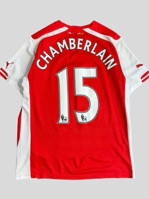 Arsenal matchtröja Puma röd/vit - Säljer en röd och vit Arsenal matchtröja från Puma med tryck 'Chamberlain 15' på ryggen. Tröjan har klubbmärke och Fly Emirates-logga på bröstet samt korta ärmar. Perfekt för fotbollsfans som vill visa sitt stöd för laget.  OBS FINNS PÅ FLER SIDOR, skriv innan köp