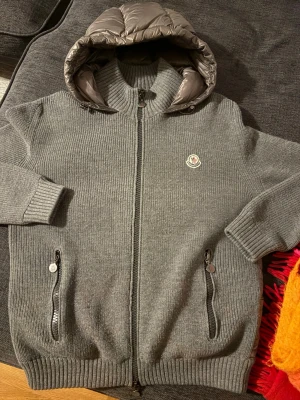 Grå Moncler cardiganjacka med huva - Snygg grå cardiganjacka från Moncler med stickad utsida och quiltad, glansig huva och insida. Jackan har dragkedja framtill, två fickor med dragkedja och Moncler-logga på bröstet. Perfekt för lager-på-lager och streetstyle.