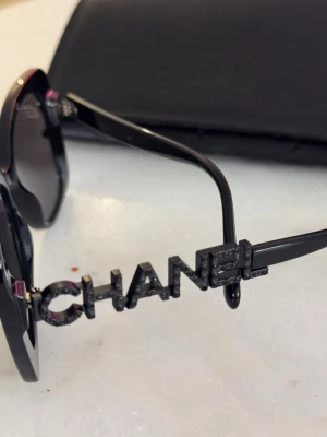 Svarta solglasögon från Chanel - Stilrena svarta solglasögon från Chanel med fyrkantig form och breda bågar. Sidan pryds av CHANEL-loggan i svart med glittrig effekt. Glasen är mörka och bågarna är i blank plast. Perfekt accessoar för en trendig look. Kvitto medföljer såklart!