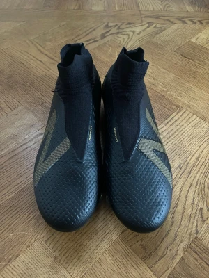 Svarta New Balance fotbollsskor - Snygga svarta fotbollsskor från New Balance med guldiga detaljer och stor N-logga på sidan. Skorna har strumpliknande skaft i textil och ovandel i syntetmaterial med struktur för bättre bollkontroll. Perfekta för dig som vill sticka ut på planen. Använda en gång, nyskick! 